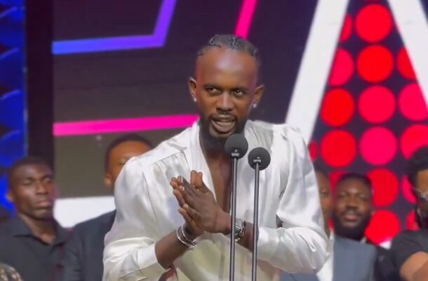 Man in a white shirt speaks into a microphone on a brightly lit stage, hands pressed together in front of him.