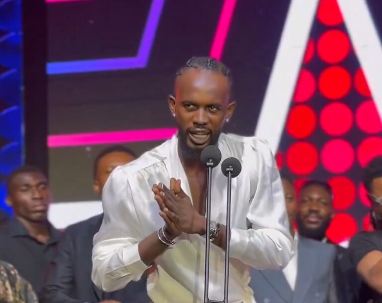Man in a white shirt speaks into a microphone on a brightly lit stage, hands pressed together in front of him.
