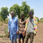 Three men with blurred faces stand in a dry, rural area next to a tarp-covered pile of a firearm and bullets laid on the ground.