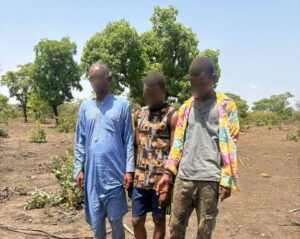 Three men with blurred faces stand in a dry, rural area next to a tarp-covered pile of a firearm and bullets laid on the ground.