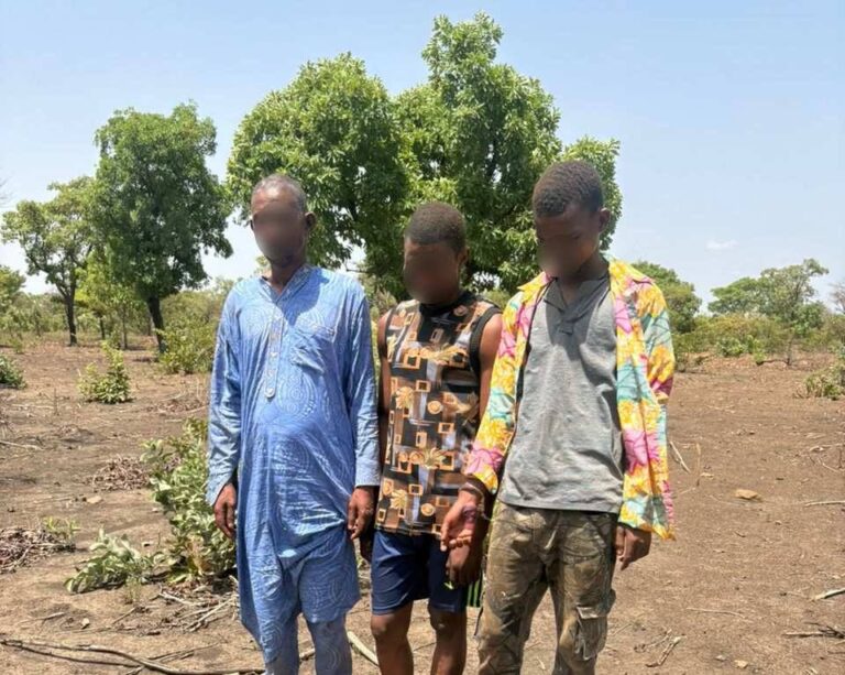 Three men with blurred faces stand in a dry, rural area next to a tarp-covered pile of a firearm and bullets laid on the ground.