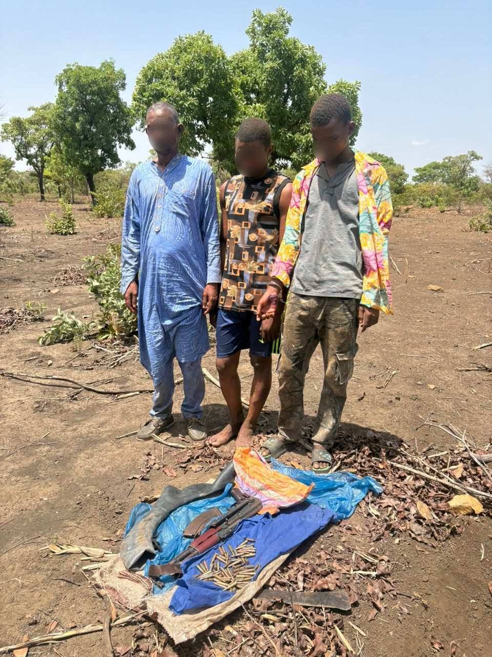 Three men with blurred faces stand in a dry, rural area next to a tarp-covered pile of a firearm and bullets laid on the ground.