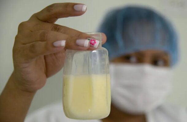Healthcare worker in mask and cap holds a small bottle of pale yellow liquid toward the camera.