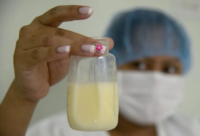 Healthcare worker in mask and cap holds a small bottle of pale yellow liquid toward the camera.