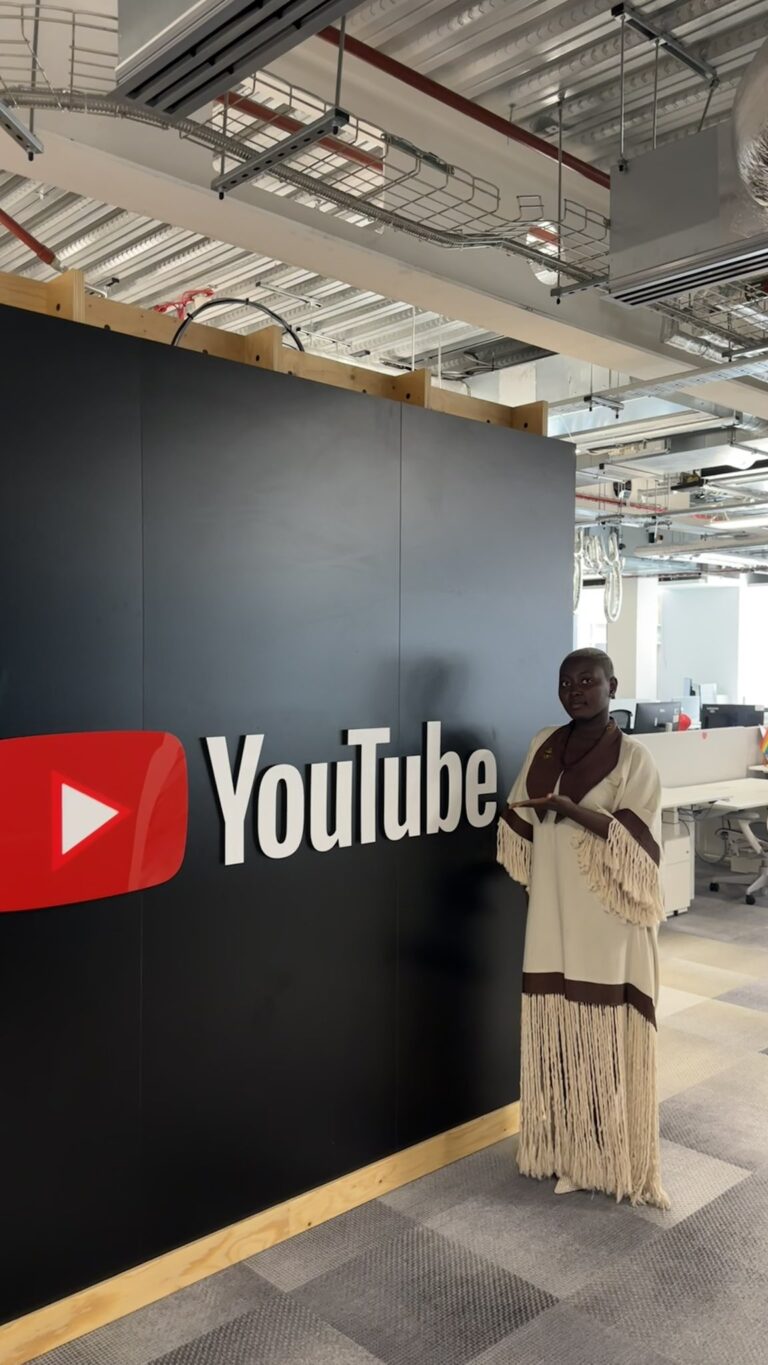 Woman in a cream and brown fringed dress stands beside a large YouTube logo on a black wall in an office setting.