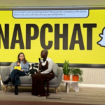 Two people sit on a sofa on stage with a huge Snapchat sign behind them, speaking into microphones.
