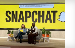 Two people sit on a sofa on stage with a huge Snapchat sign behind them, speaking into microphones.