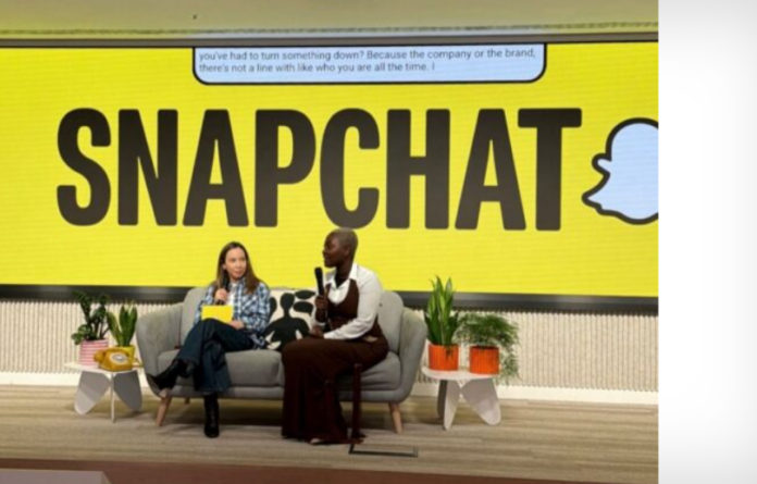 Two people sit on a sofa on stage with a huge Snapchat sign behind them, speaking into microphones.