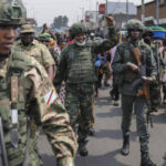 Armed soldiers in camouflage uniforms march down a street as civilians watch a crowd around them.