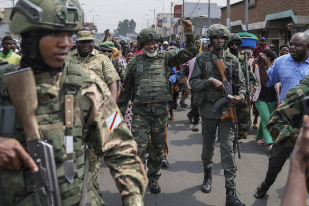 Armed soldiers in camouflage uniforms march down a street as civilians watch a crowd around them.