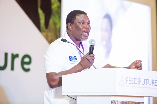 Man in a white polo shirt speaks into a microphone at a podium with FEED THE FUTURE branding and a large screen behind him