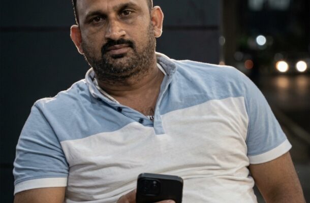 Man in a light blue and white polo shirt holds a smartphone outdoors at night.
