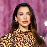 Model walking a fashion show wearing a yellow and black leopard‑print top against a pink blurred runway backdrop.