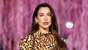Model walking a fashion show wearing a yellow and black leopard‑print top against a pink blurred runway backdrop.