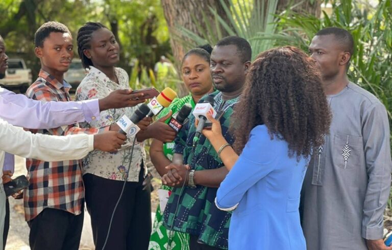 Man being interviewed by multiple reporters outdoors, microphones aimed at him as cameras and journalists surround him.