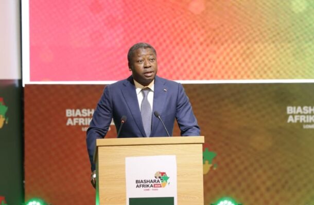 Faure-Essozimna-Gnasisingbe2 | Ghanaguardian.com Man in a navy suit speaks at a wooden podium on a colorful stage, Biashara Afrika branding visible in the background.