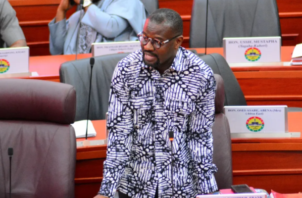 Man in a black-and-white patterned shirt standing at a conference table with a microphone, in a formal meeting room.