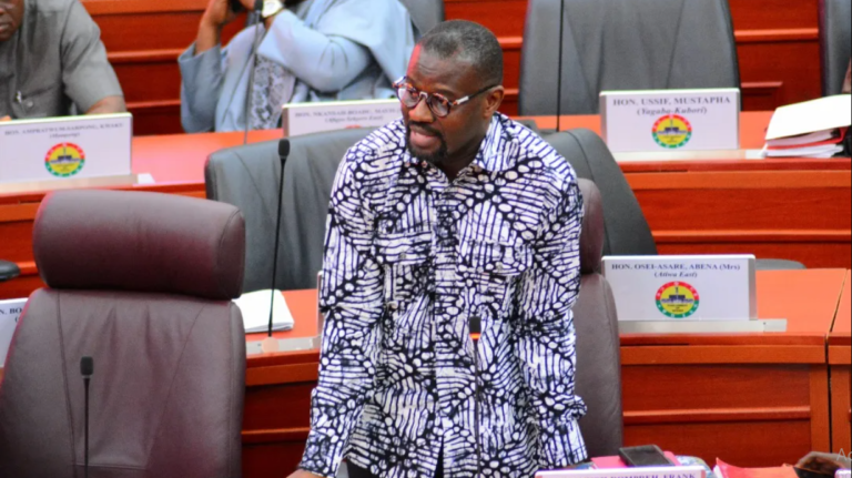 Man in a black-and-white patterned shirt standing at a conference table with a microphone, in a formal meeting room.