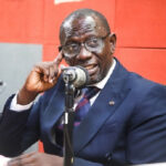Man in a navy suit and glasses speaks into a microphone in a radio studio with a red backdrop.