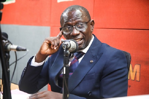 Man in a navy suit and glasses speaks into a microphone in a radio studio with a red backdrop.