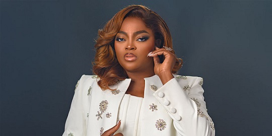 Portrait of a confident woman in a white embroidered blazer posing against a dark blue background.