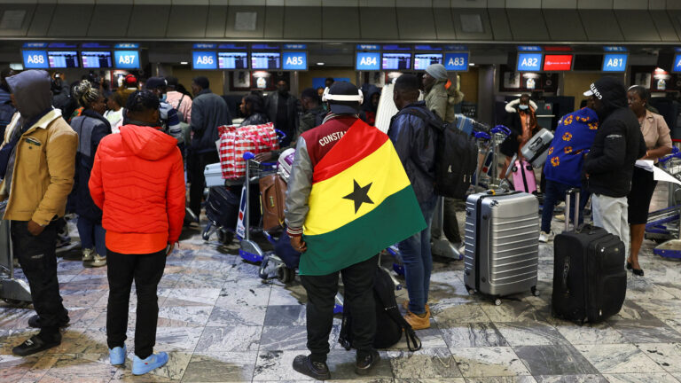 Crowded airport check-in area with travelers and rolling luggage; a man in the center wears the Senegal flag as a cape.