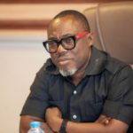 Man in a black short-sleeve shirt seated at a conference table, wearing black-framed glasses with red temples and a serious expression; watermark at lower-left says Ghana Shippers Authority.