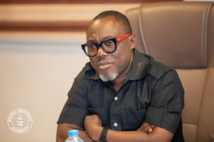 Man in a black short-sleeve shirt seated at a conference table, wearing black-framed glasses with red temples and a serious expression; watermark at lower-left says Ghana Shippers Authority.