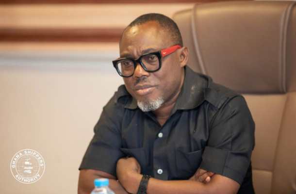 Man in a black short-sleeve shirt seated at a conference table, wearing black-framed glasses with red temples and a serious expression; watermark at lower-left says Ghana Shippers Authority.
