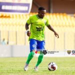 Soccer player in a neon green jersey and blue shorts dribbles a ball on a grass field, with yellow stadium seats in the background.