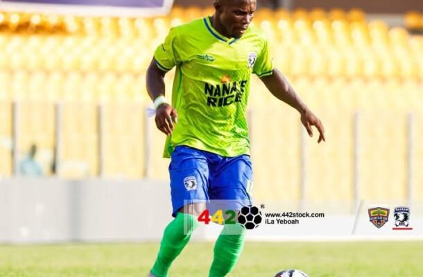 Soccer player in a neon green jersey and blue shorts dribbles a ball on a grass field, with yellow stadium seats in the background.