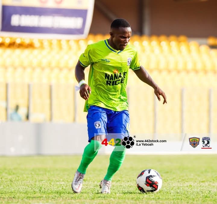 Soccer player in a neon green jersey and blue shorts dribbles a ball on a grass field, with yellow stadium seats in the background.