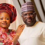 A smiling couple in traditional Nigerian attire posing indoors: the woman wears a pink patterned dress and a large red gele, while the man wears a cream kaftan and patterned cap.