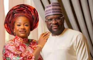 A smiling couple in traditional Nigerian attire posing indoors: the woman wears a pink patterned dress and a large red gele, while the man wears a cream kaftan and patterned cap.