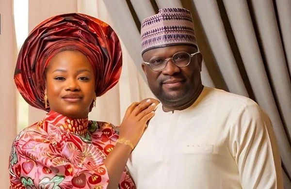 Portrait of a couple in traditional Nigerian attire: woman in a vibrant red gele and floral dress, man in a cream agbada and cap, posing together indoors.
