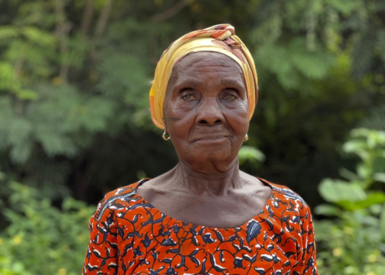 Portrait of an elderly African woman wearing a yellow head wrap and an orange patterned dress in a lush garden.