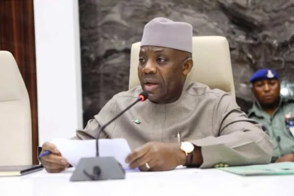 Man in a gray traditional outfit and cap speaks into a microphone at a conference table, holding papers; security personnel in the background.