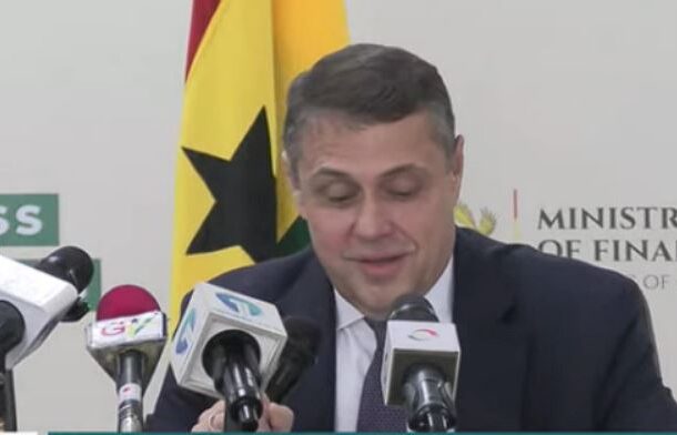 Man in a suit at a press conference with multiple microphones, flag and Ministry of Finance backdrop behind him, speaking to the media in Ghanaian setting