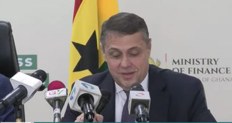 Man in a suit at a press conference with multiple microphones, flag and Ministry of Finance backdrop behind him, speaking to the media in Ghanaian setting