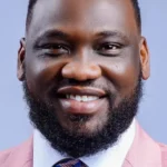 Portrait of a smiling man with a beard in a pink suit jacket and white shirt against a blue background.