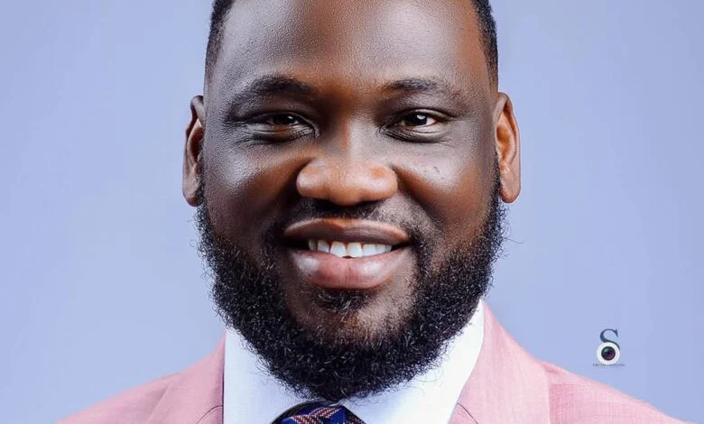 Portrait of a smiling man with a beard in a pink suit jacket and white shirt against a blue background.
