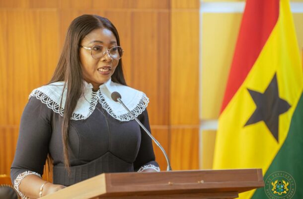 Female speaker at a wooden podium delivering a speech, wearing a black dress with a white lace collar and glasses, microphone in front of her.