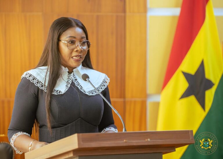 Female speaker at a wooden podium delivering a speech, wearing a black dress with a white lace collar and glasses, microphone in front of her.