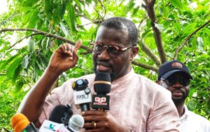 Man wearing glasses and a pink shirt speaks to reporters outdoors, with microphones in front of him.