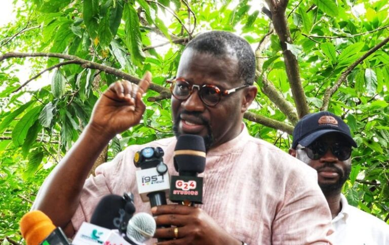 Man wearing glasses and a pink shirt speaks to reporters outdoors, with microphones in front of him.