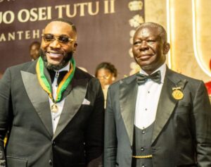 Two men in black tuxedos at a formal event, one wearing a gold medal on a green-yellow ribbon beside the other in a bow tie and pin image backdrop behind them
