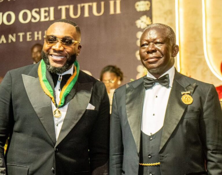 Two men in black tuxedos at a formal event, one wearing a gold medal on a green-yellow ribbon beside the other in a bow tie and pin image backdrop behind them