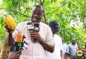 Man in a pink shirt holds a cacao pod while speaking into multiple microphones at an outdoor press briefing, with reporters nearby and a tree-filled background.