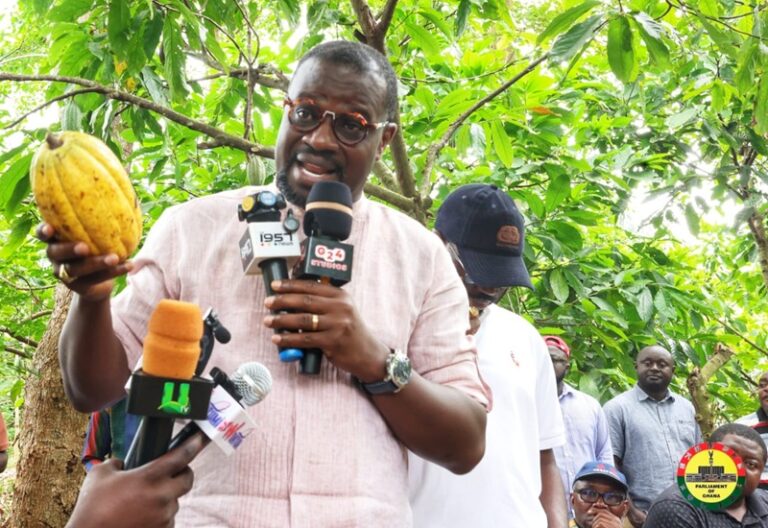 Man in a pink shirt holds a cacao pod while speaking into multiple microphones at an outdoor press briefing, with reporters nearby and a tree-filled background.