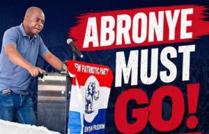 Man speaks at a political rally from behind a podium; a large sign behind him reads 'ABRONYE MUST GO!' in bold white and red letters.